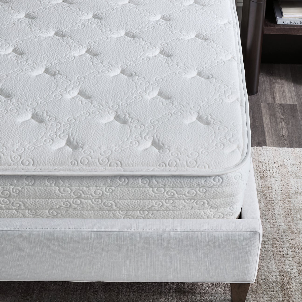 A close-up of a Luxury Sferra Sognofacile Mattress in a Box with white patterns on a bed frame beside a nightstand on a wood floor.