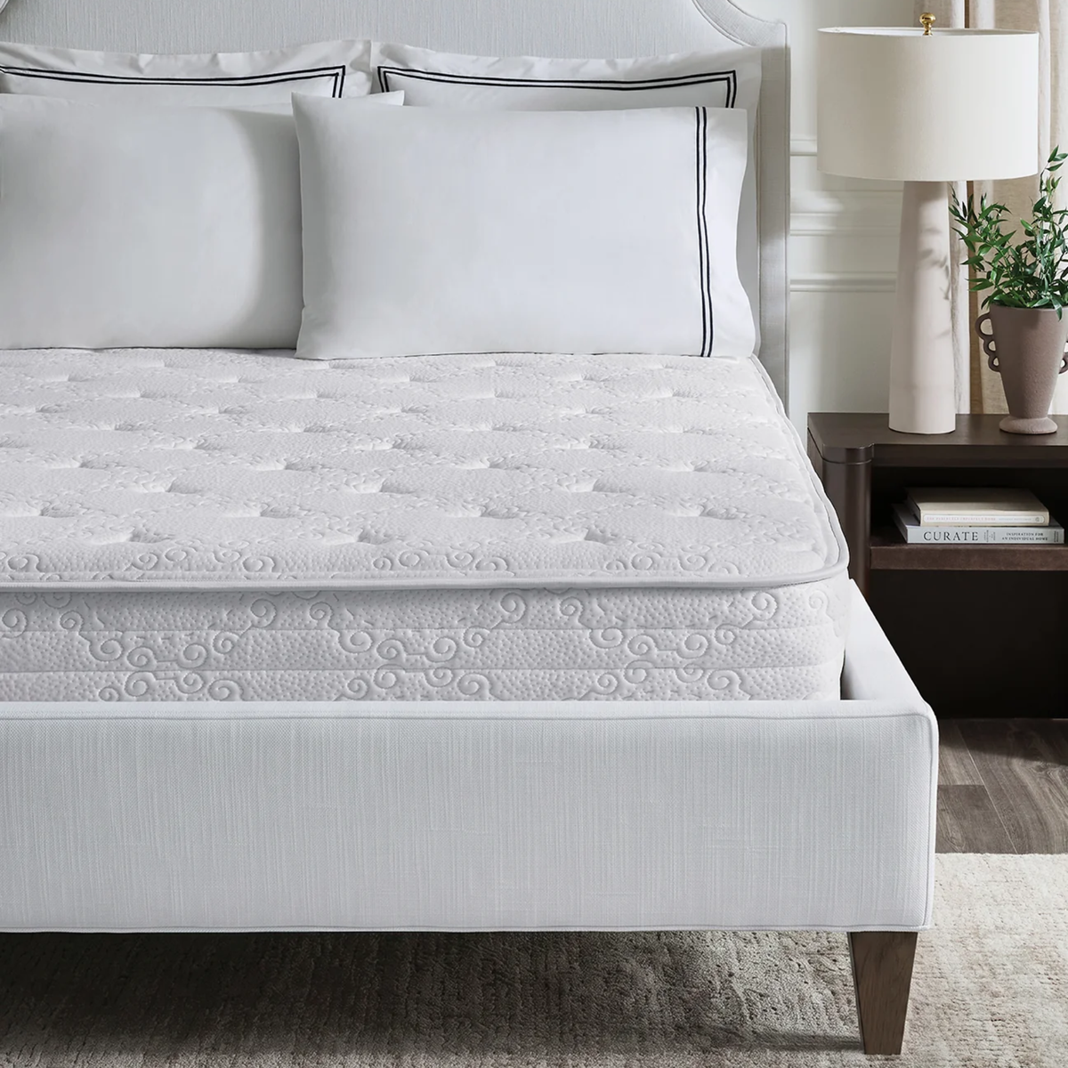 A neatly made bed with white bedding and pillows showcases a Sferra Sognofacile Mattress in a Box beside a nightstand with a lamp.