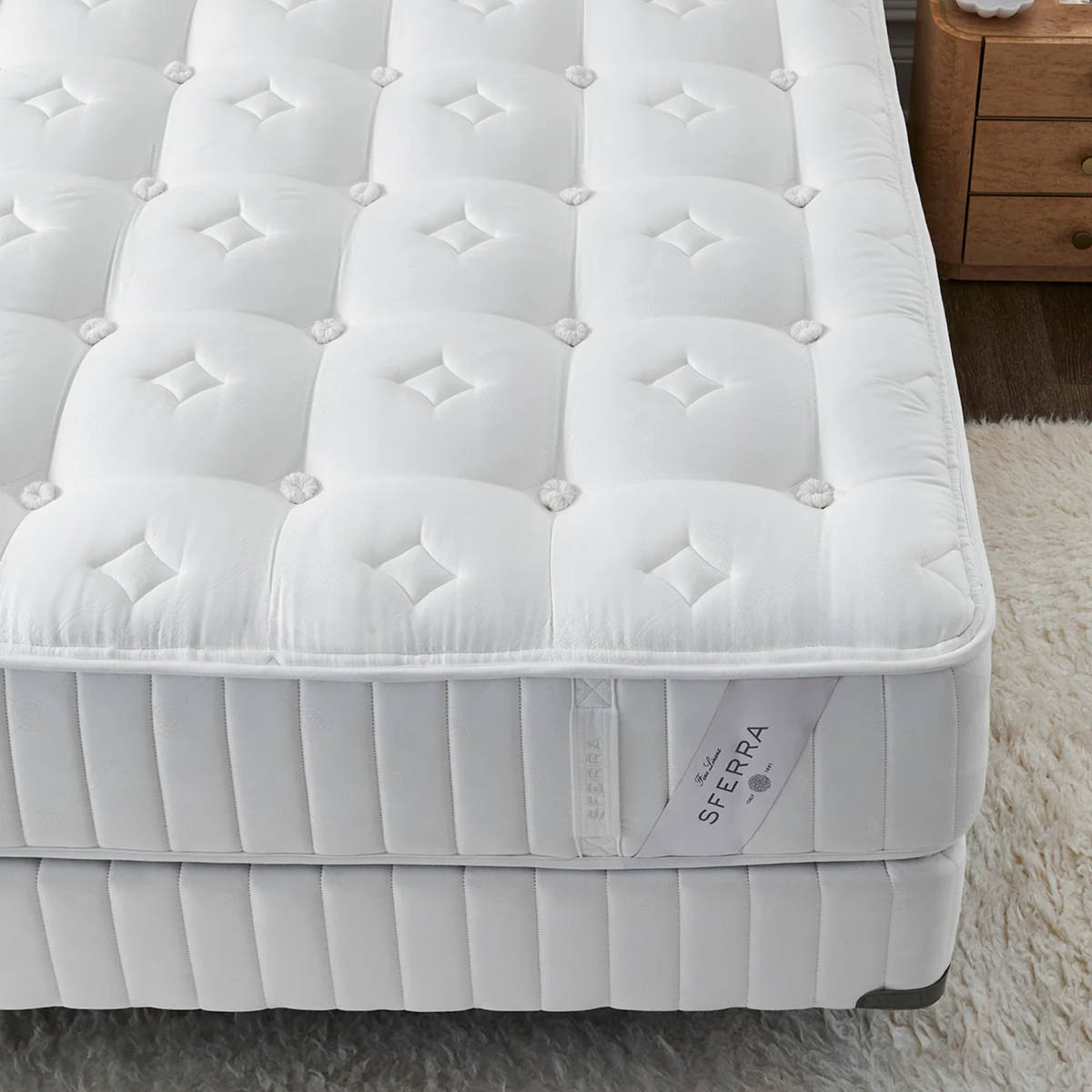 White, tufted Luxury Sferra Sonno Notte CV Luxury Firm Mattress on box spring next to nightstand and white rug, with a visible Serra brand tag.