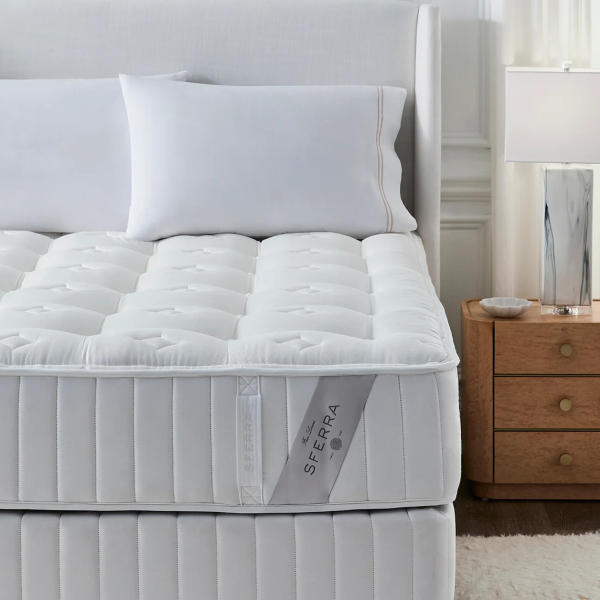 A Sferra Sonno Notte CV Luxury Firm Mattress, white pillows, and a bedside table with a lamp in a bright bedroom.