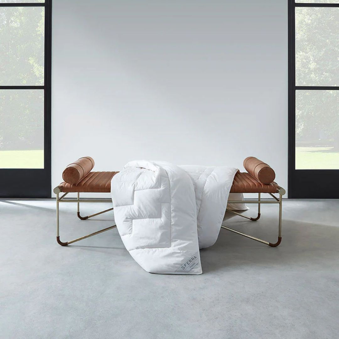 A hypoallergenic duvet draped over a brown leather bench in front of large, black-framed windows.