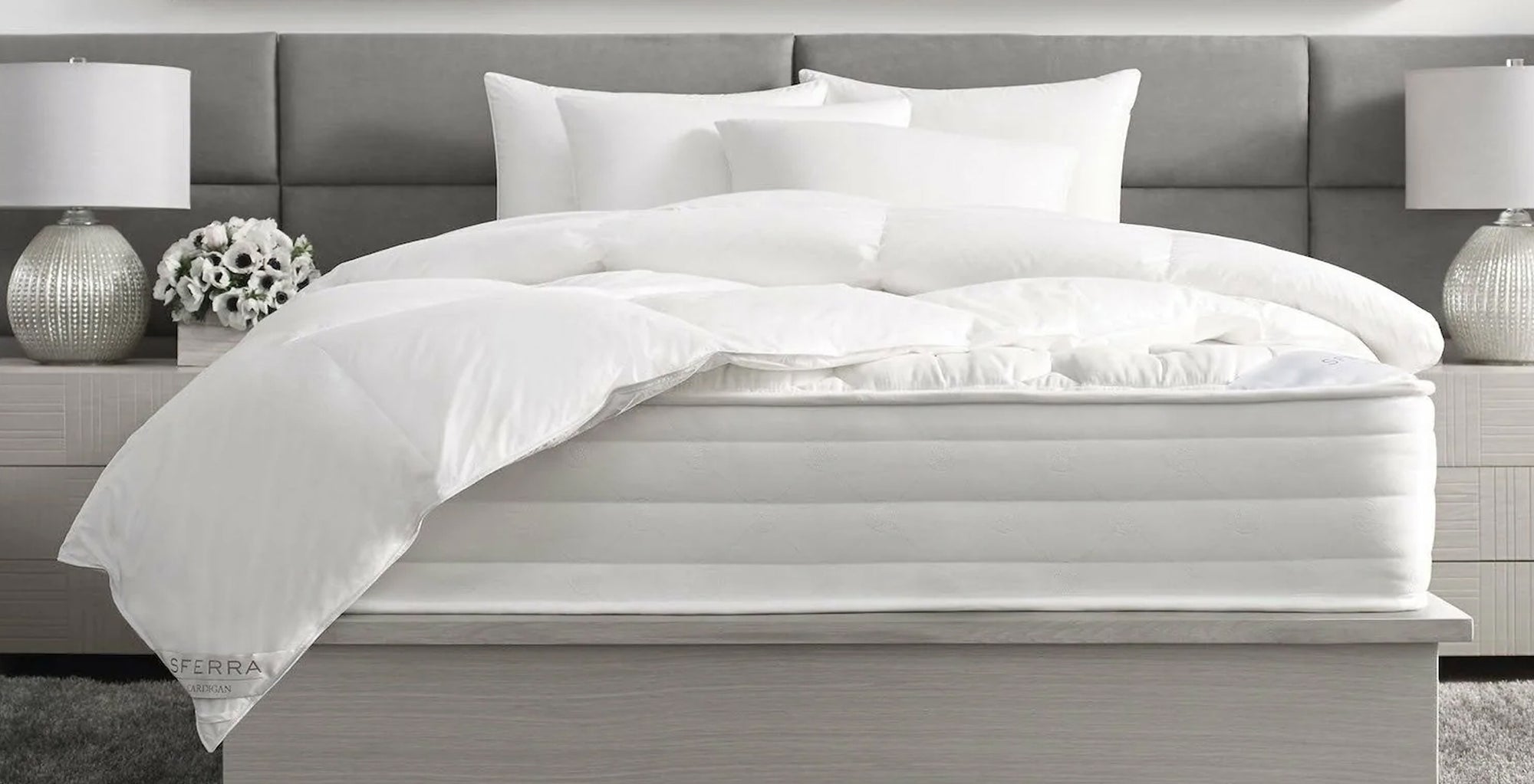 A neatly made bed with white bedding, pillows, two side lamps, and a flower vase on a modern headboard.