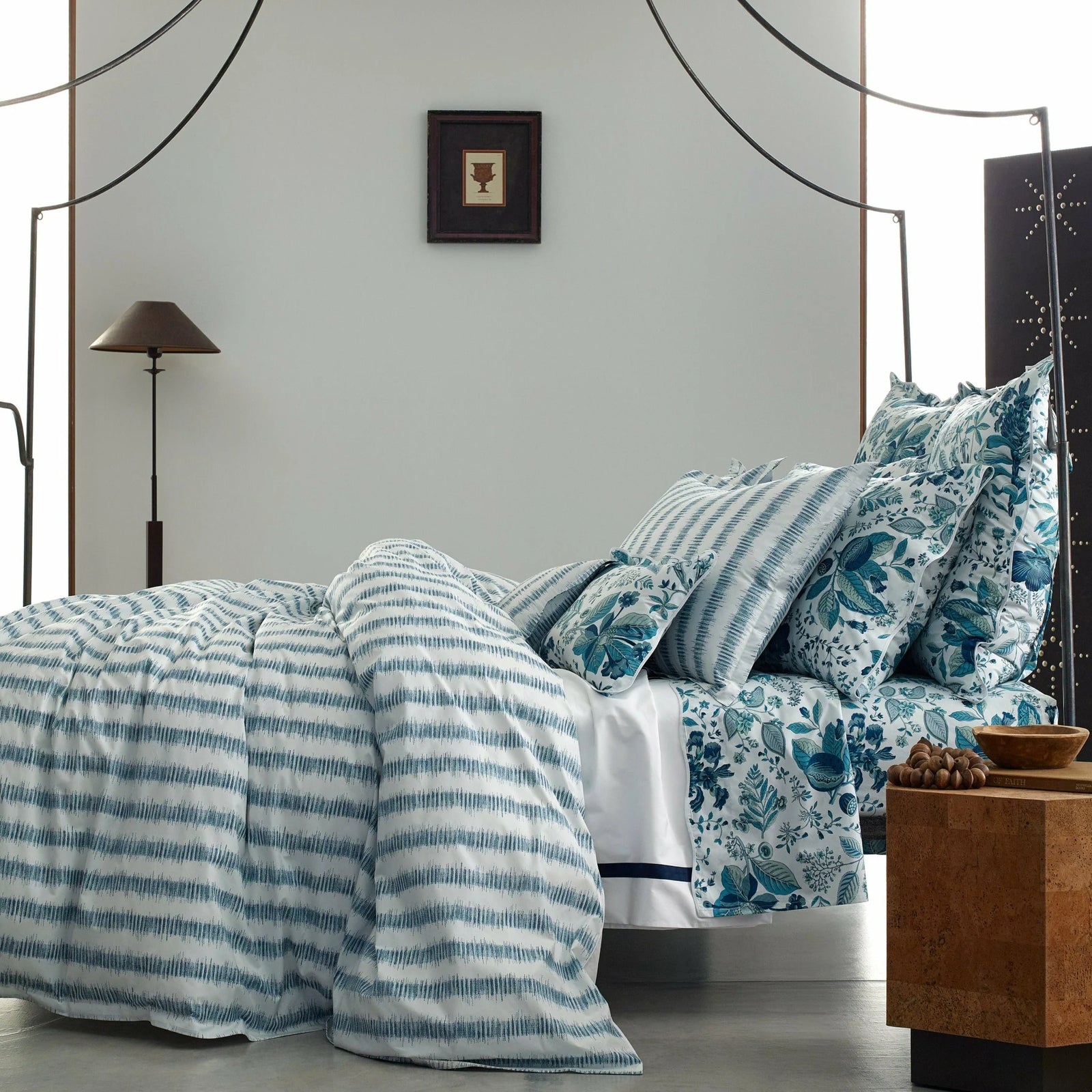 A neatly made bed with blue and white striped and floral bedding, next to a lamp, nightstand, and wall art.