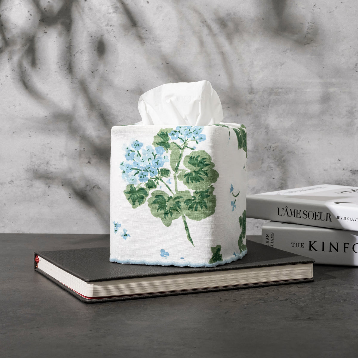 Tissue box with floral design on a stack of books against a textured wall.