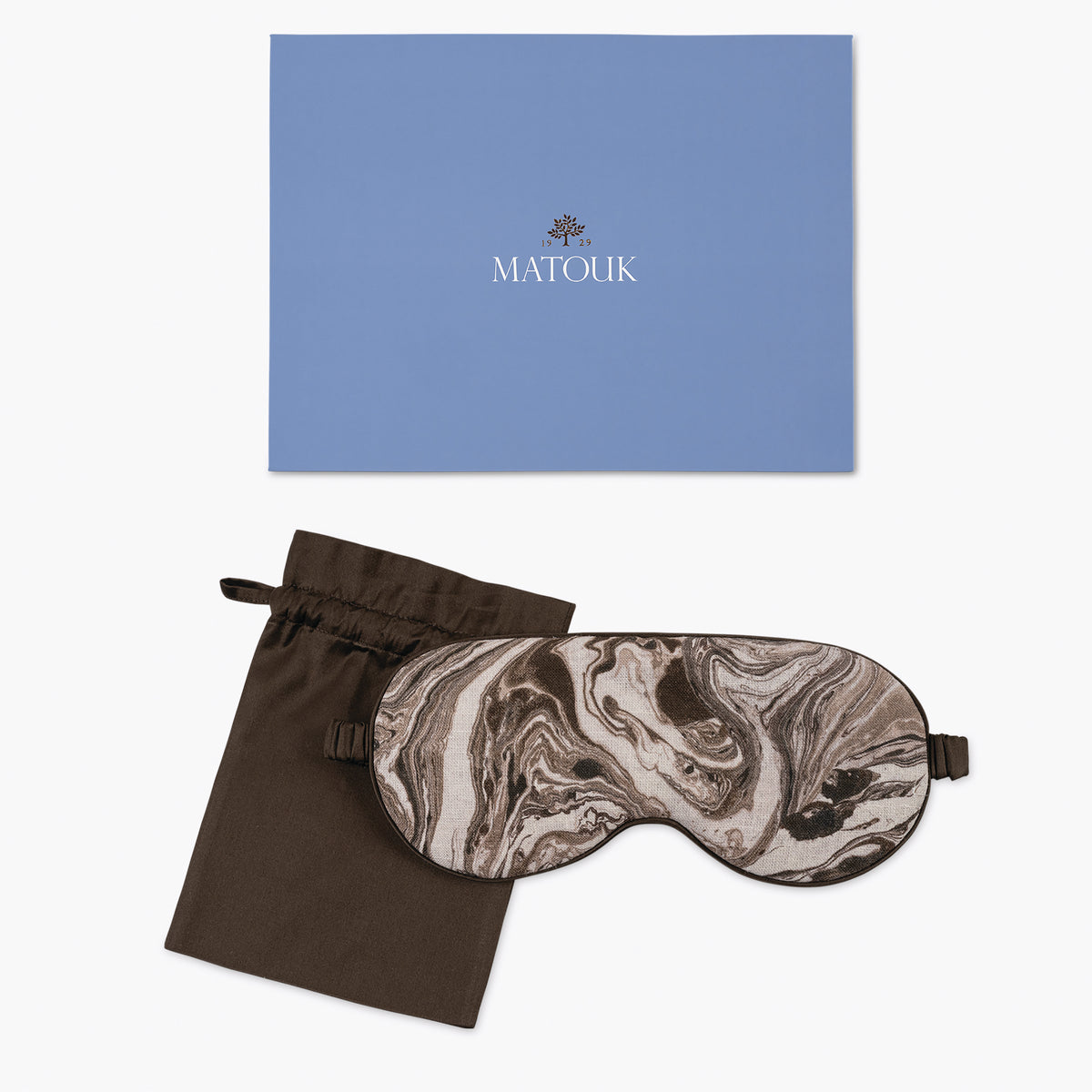 A marbled sleep mask with blackout lining and a brown pouch sits before a blue Matouk Schumacher box on white.
