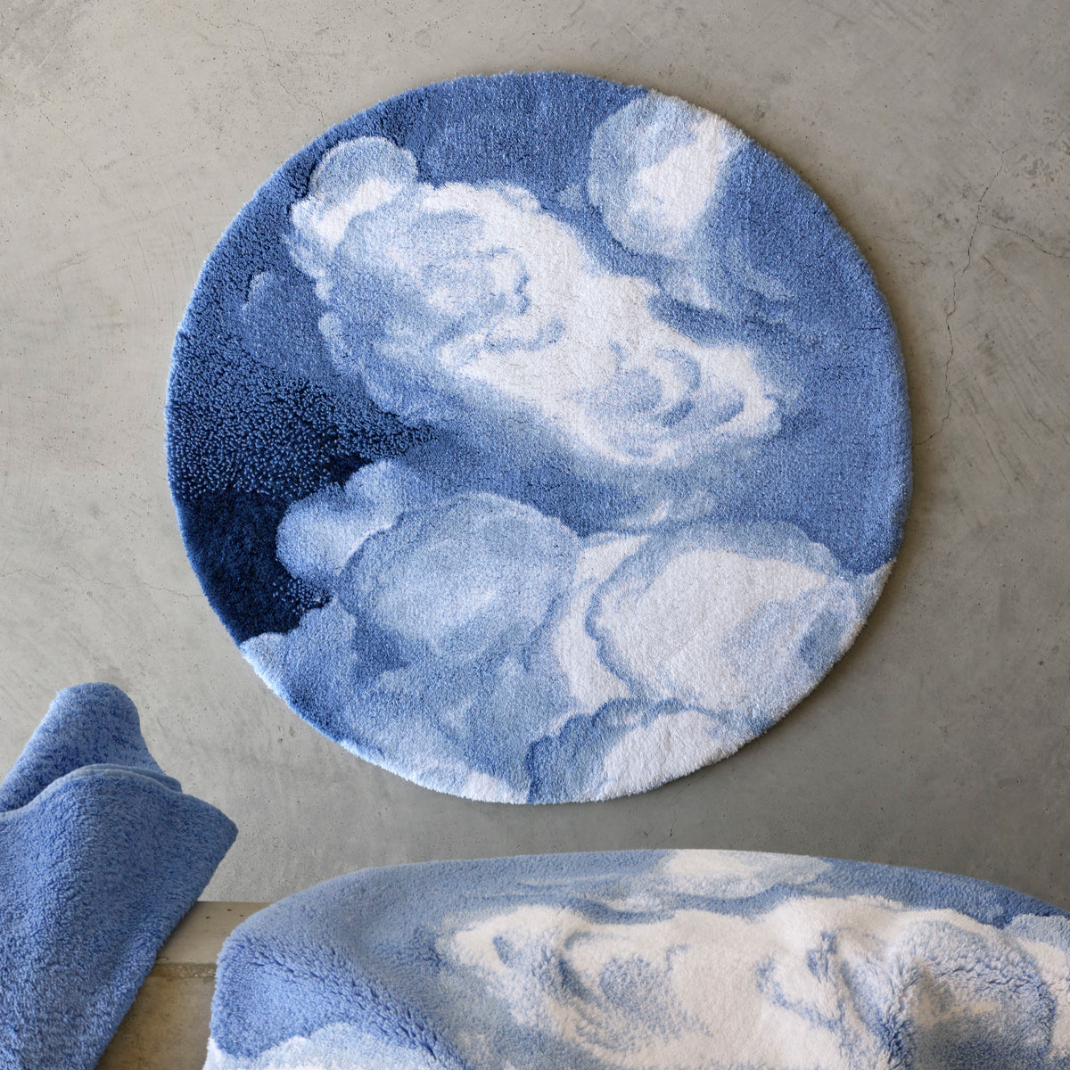 A round blue and white plush absorbent bath rug with a cloud pattern hangs on a gray wall. Matching towel and blanket nearby.