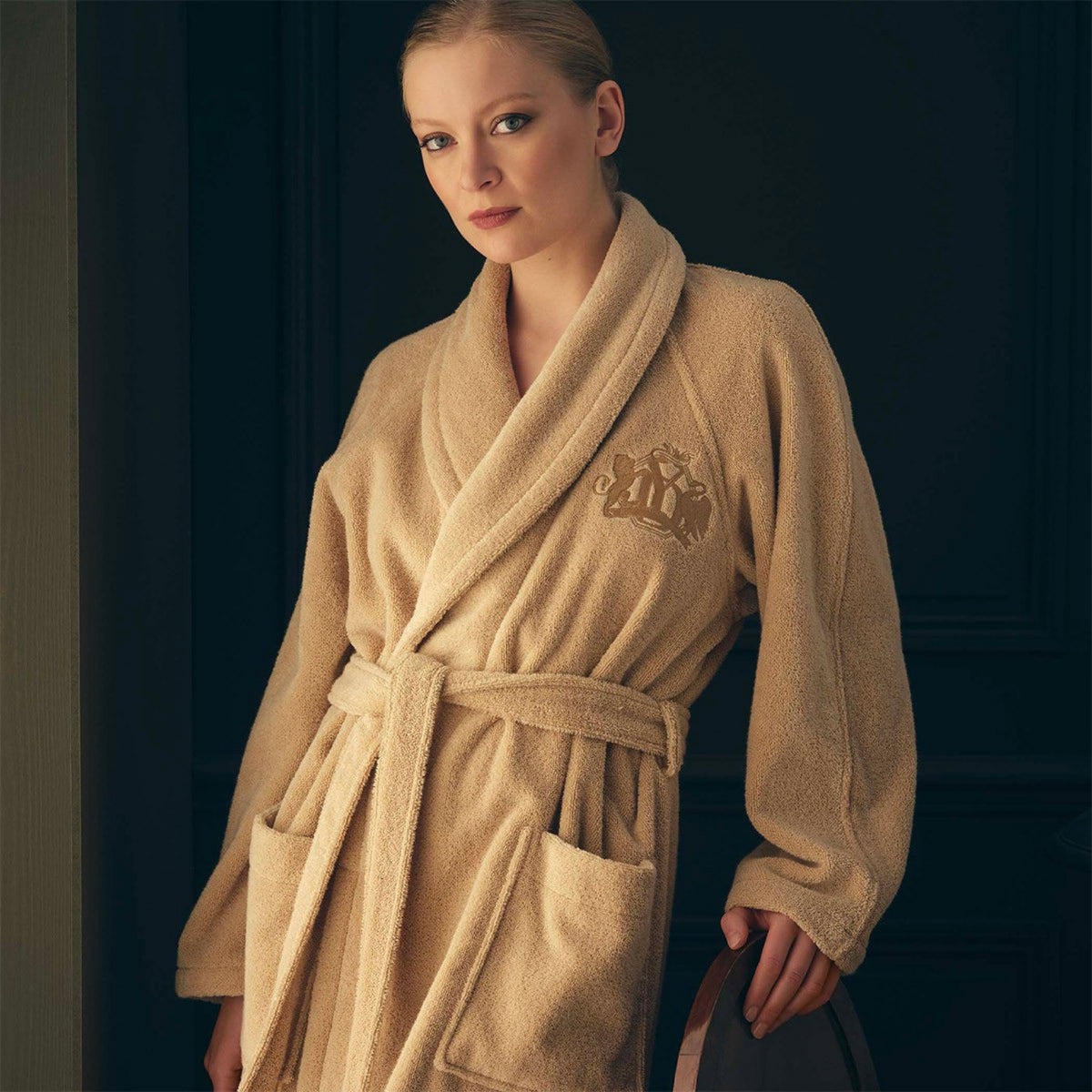 Woman in a Yves Delorme Apollon Bath Robe in Dune color made in France stands indoors, leaning on a chair, with a dark background behind her.