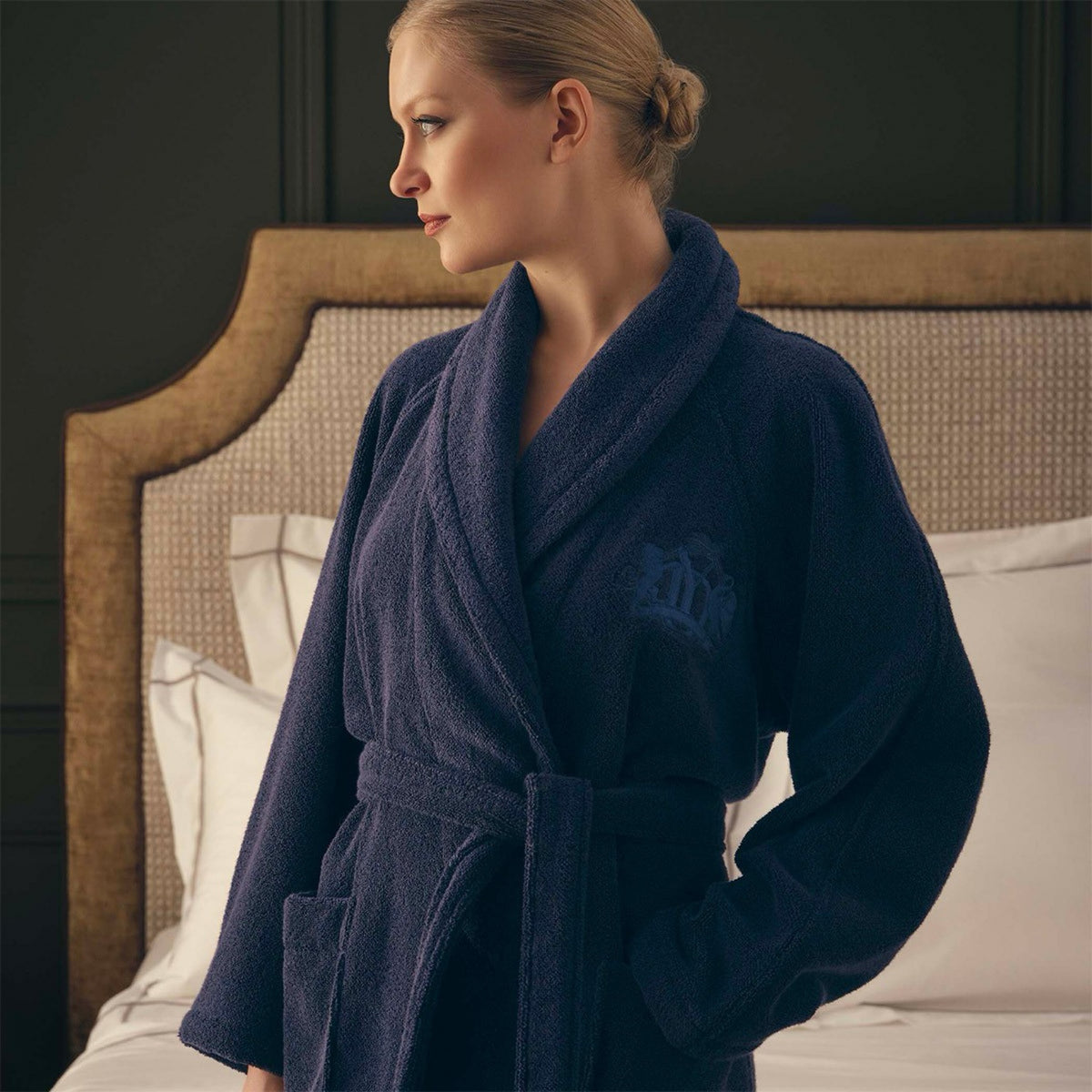 Woman in a Yves Delorme Apollon Bath Robe in Marine color made in France stands by the bed, looking left, in a softly lit room with neutral expression.