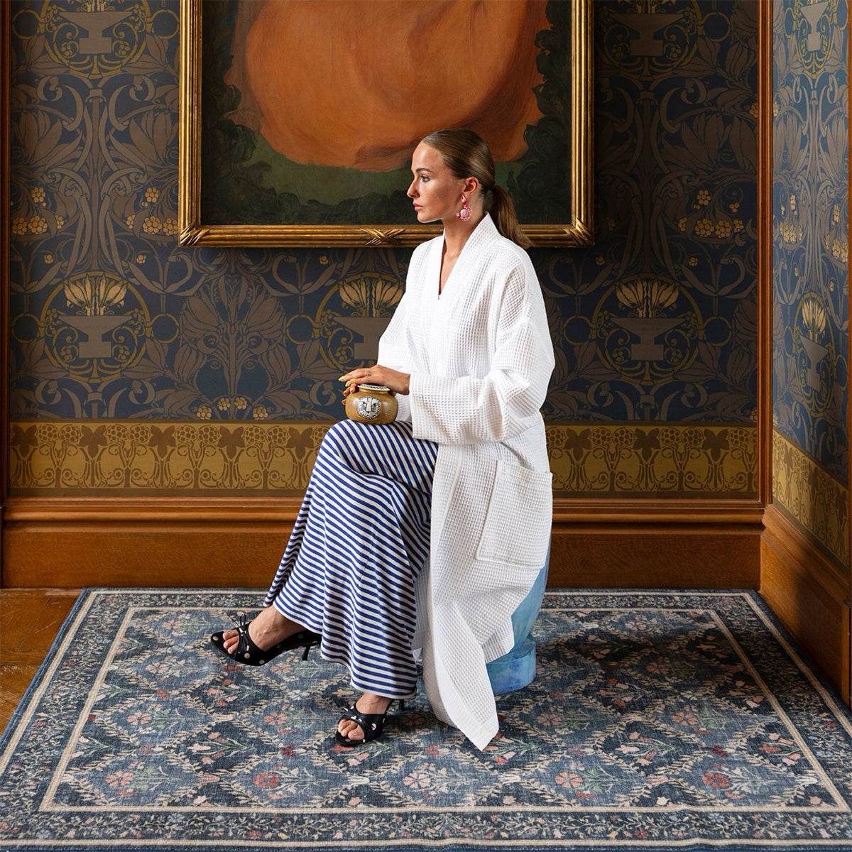 Woman in a Blanc Yves Delorme Astreena Bath Robe and striped skirt sits on a stool in a room with floral wallpaper.