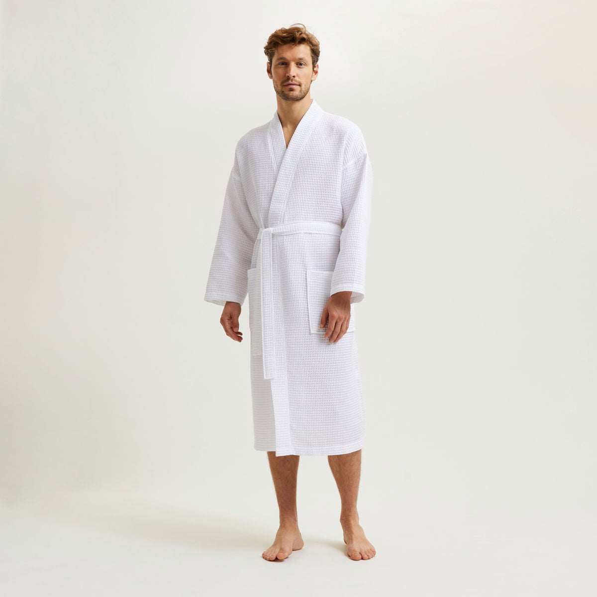 A man stands barefoot in a Blanc Yves Delorme Astreena Bath Robe against a plain light background