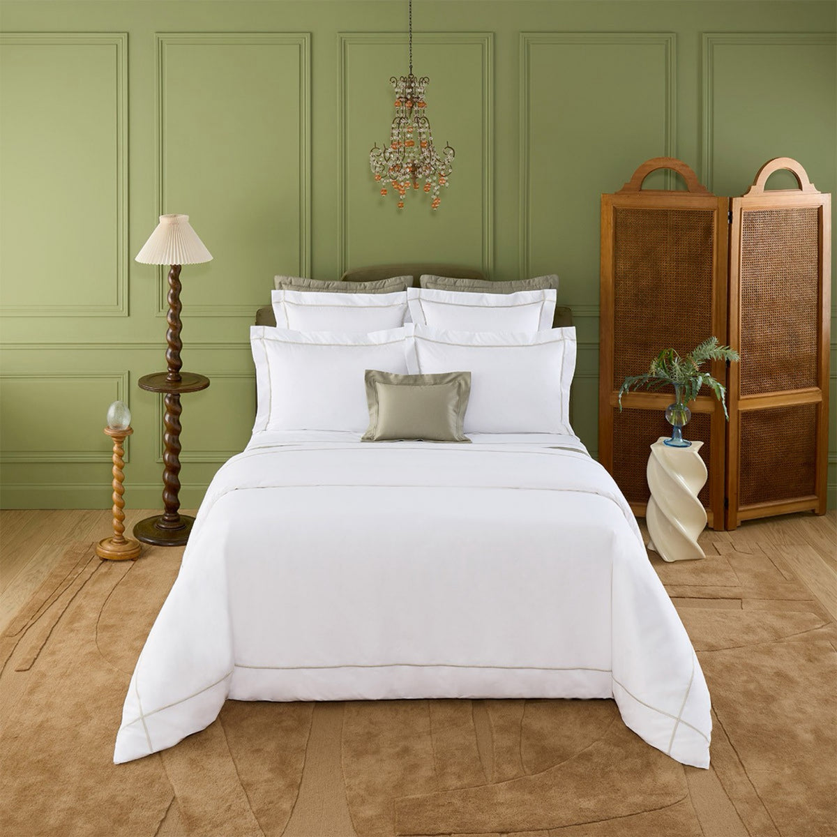 Neatly made bed with Yves Delorme Athena Bedding and Loutre embroidery, small chandelier, lamp, and accents in a green bedroom