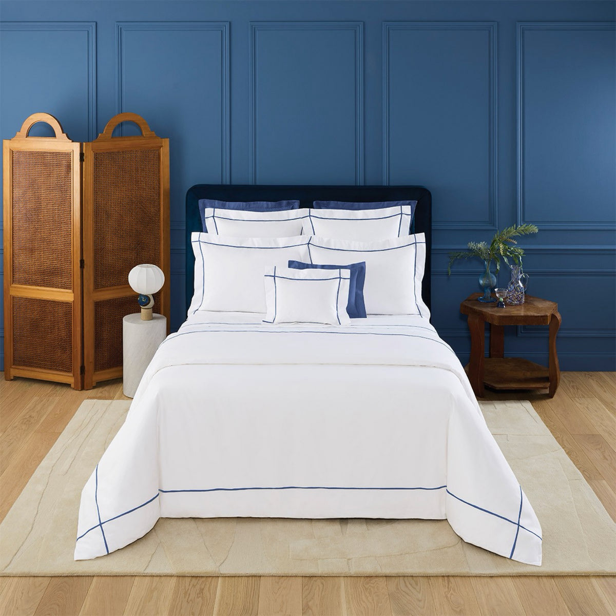 A neatly made bed with Yves Delorme Athena Bedding and Outremer embroidery, against a blue wall, with a wooden screen and side table