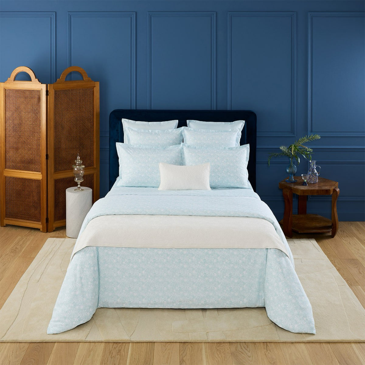 A neatly made bed with Aqua Yves Delorme Capri Bedding, set against a blue wall with wooden furniture and décor