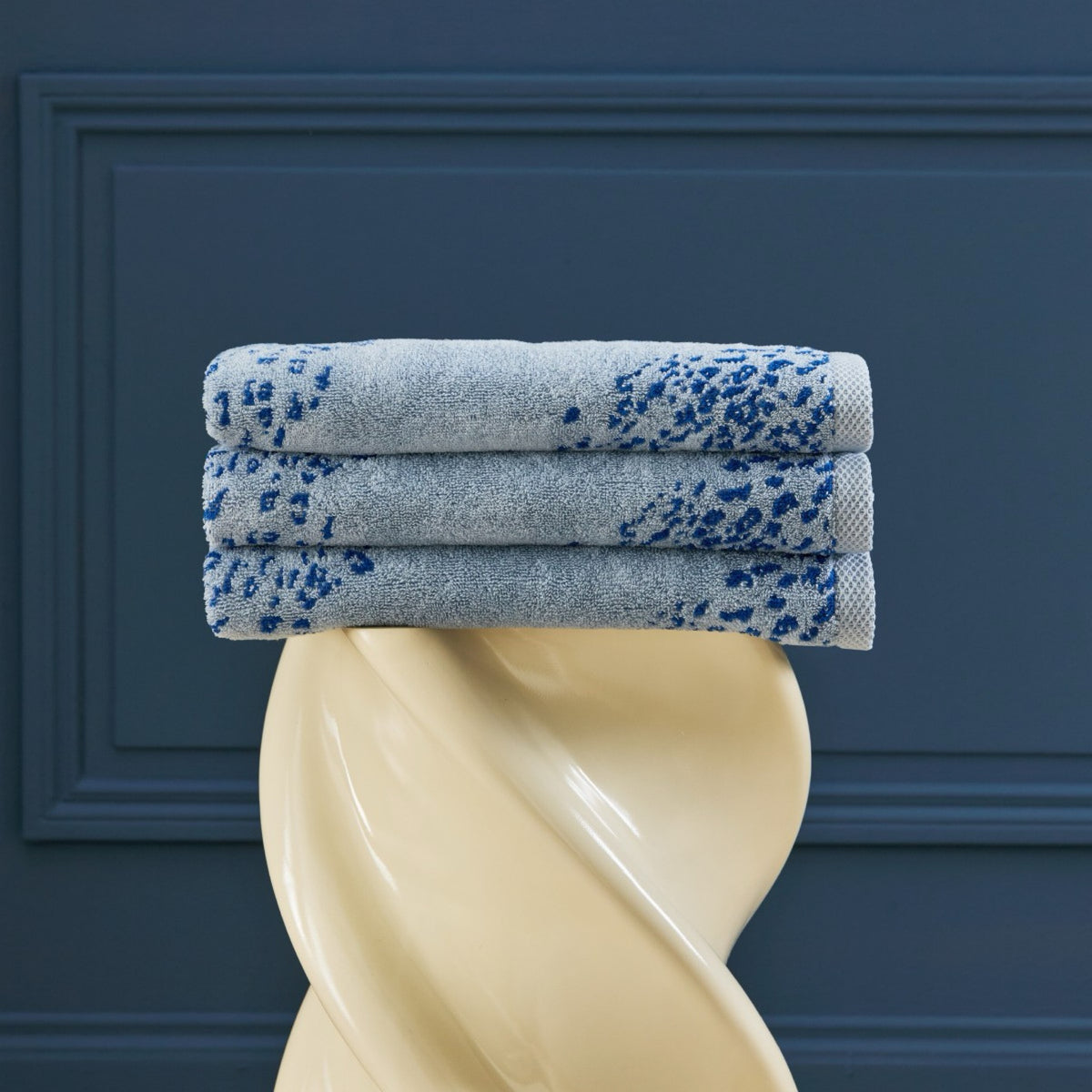 Three Yves Delorme Ecumes Bath towels are neatly stacked on a cream-colored twisted pedestal