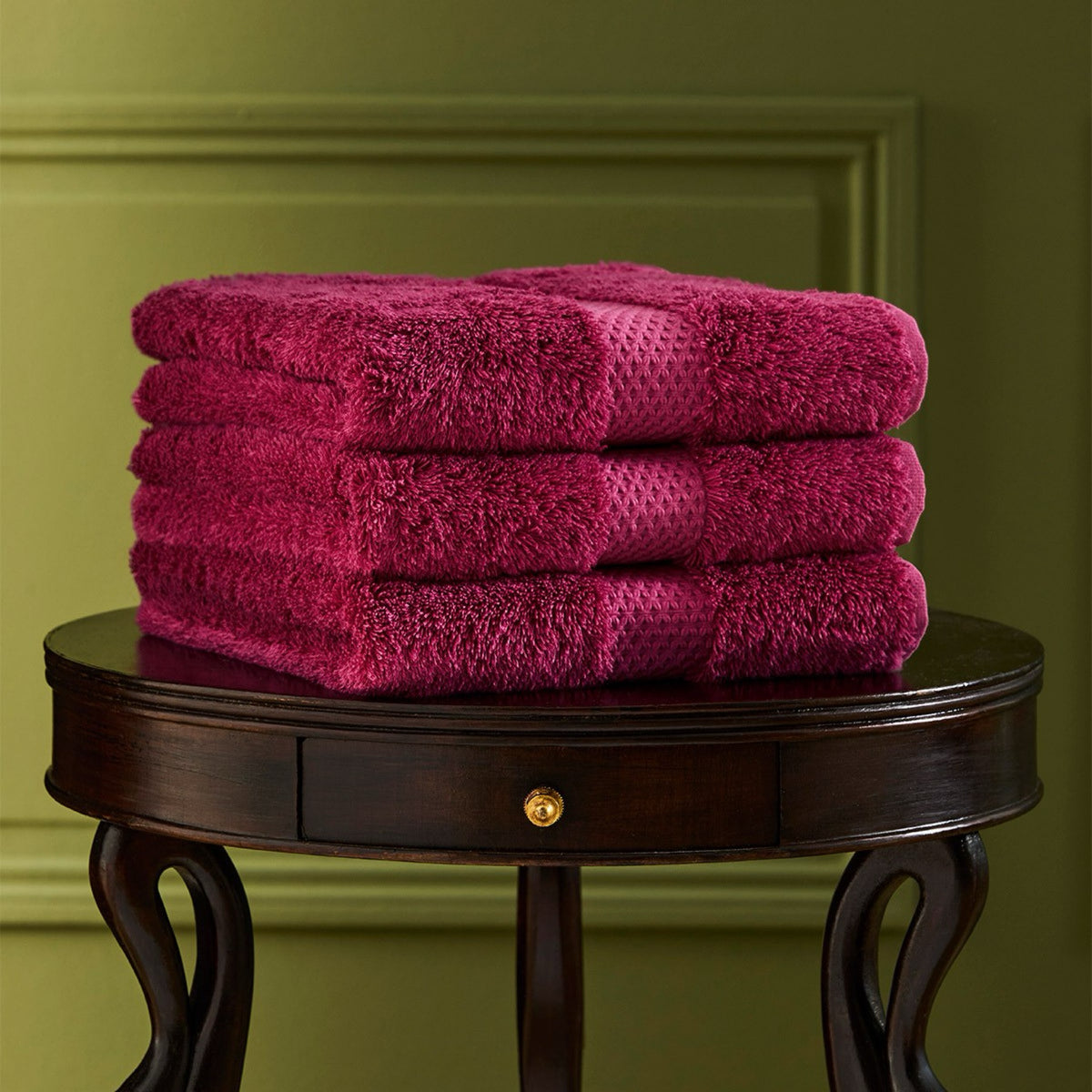 A stack of four folded Etoile Bath Towels sits on a round wooden table against a green wall.
