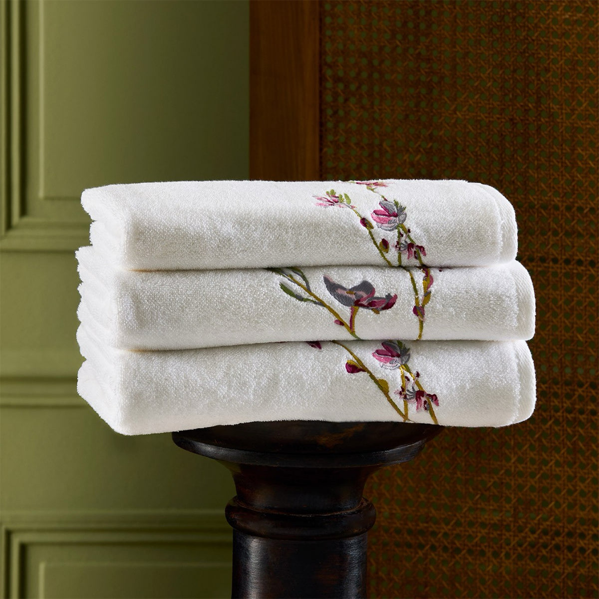 Three folded Yves Delorme Gaura Bath Towels are stacked on a dark wooden stand against a textured background