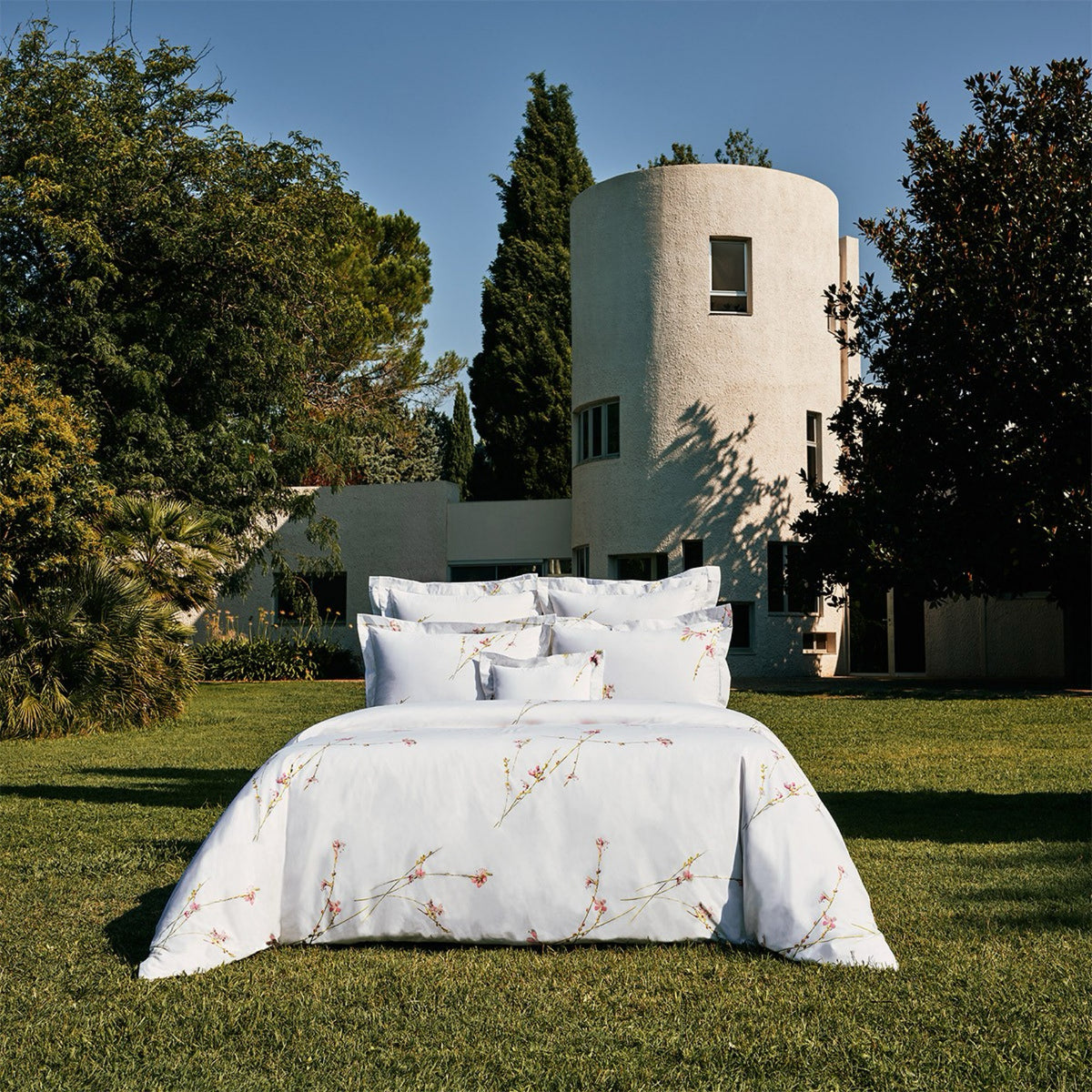 A bed with Yves Delorme Gaura Bedding Collection is placed on a lawn before a modern white cylindrical house and trees