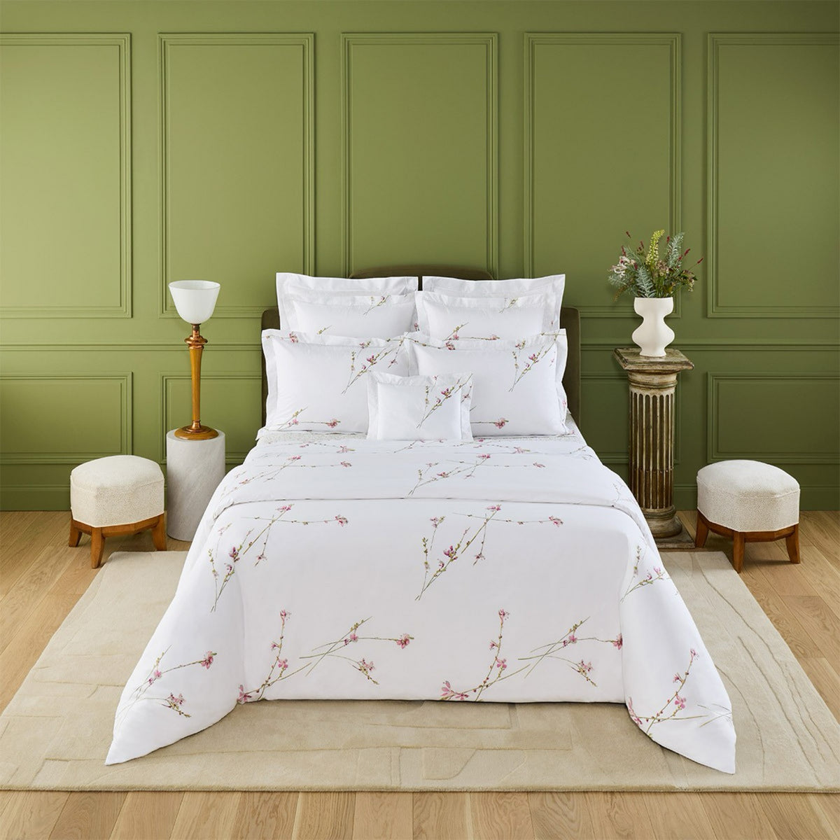A neatly made bed with Yves Delorme Gaura Bedding Collection stands against a green paneled wall, flanked by stools and tables