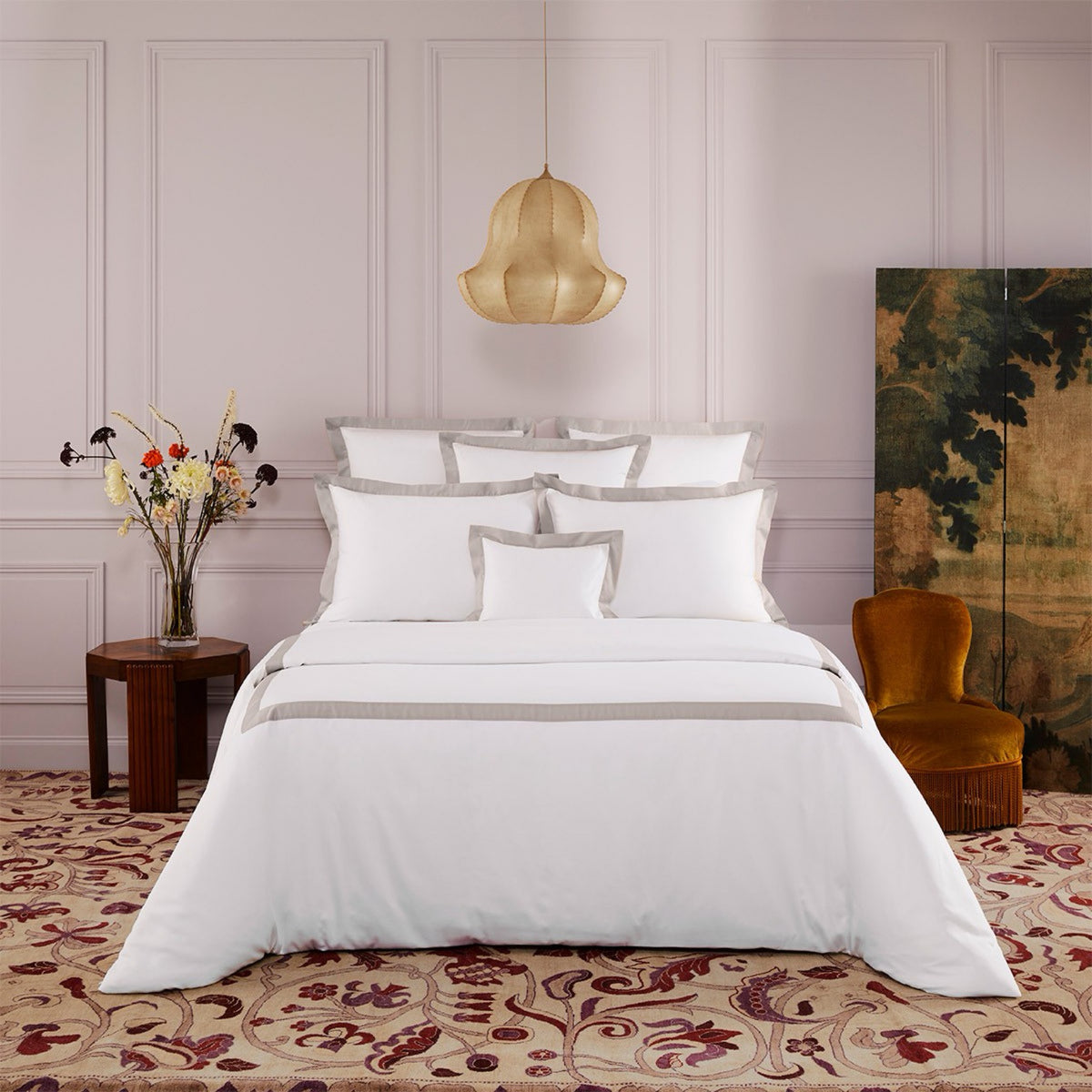 A neatly made bed Yves Delorme Linea Bedding in Loutre color, floral rug, pendant light, velvet chair, art, and flowers.