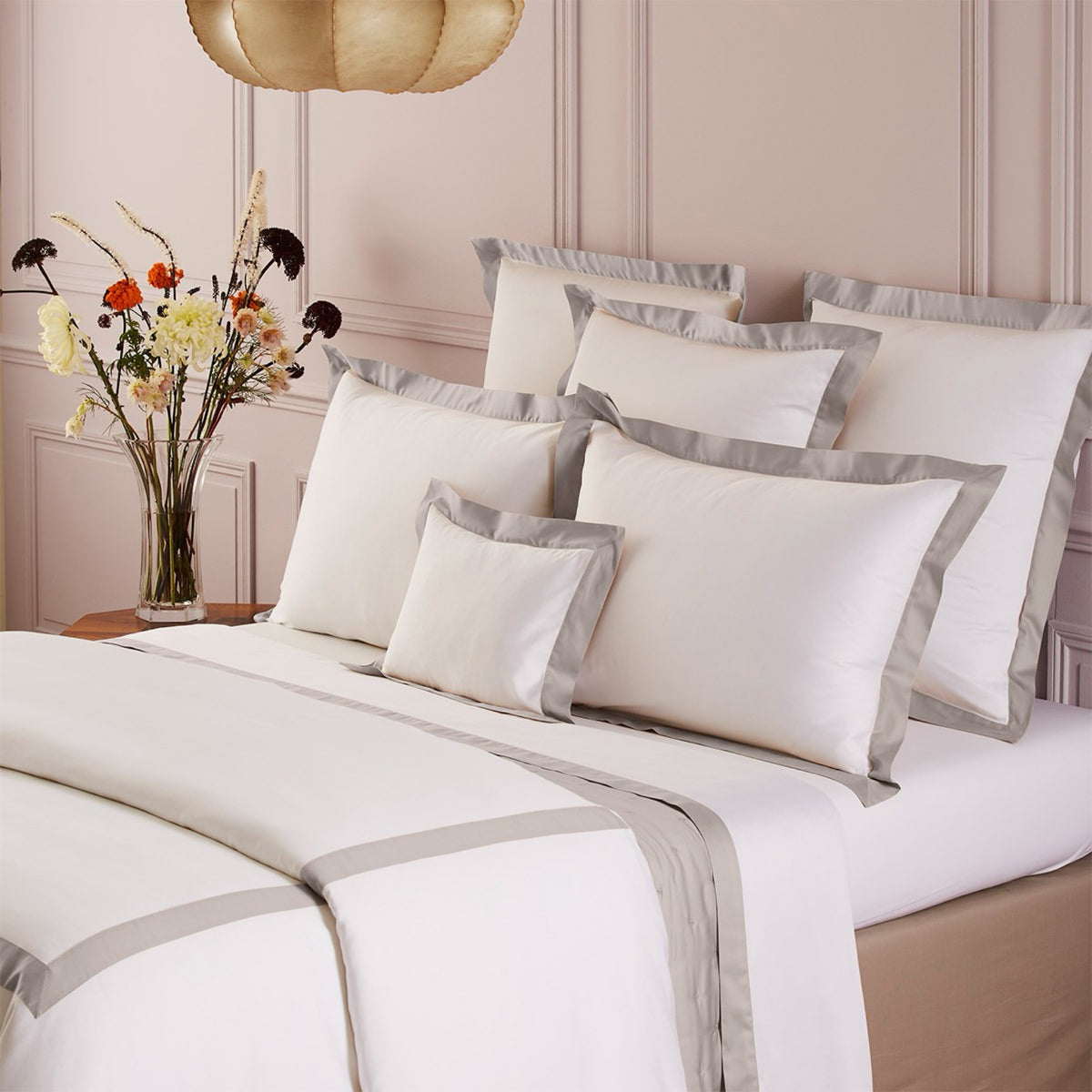 Corner view of bed with Yves Delorme Linéa cotton sateen bedding in Loutre color, beside a vase of flowers on a wooden nightstand