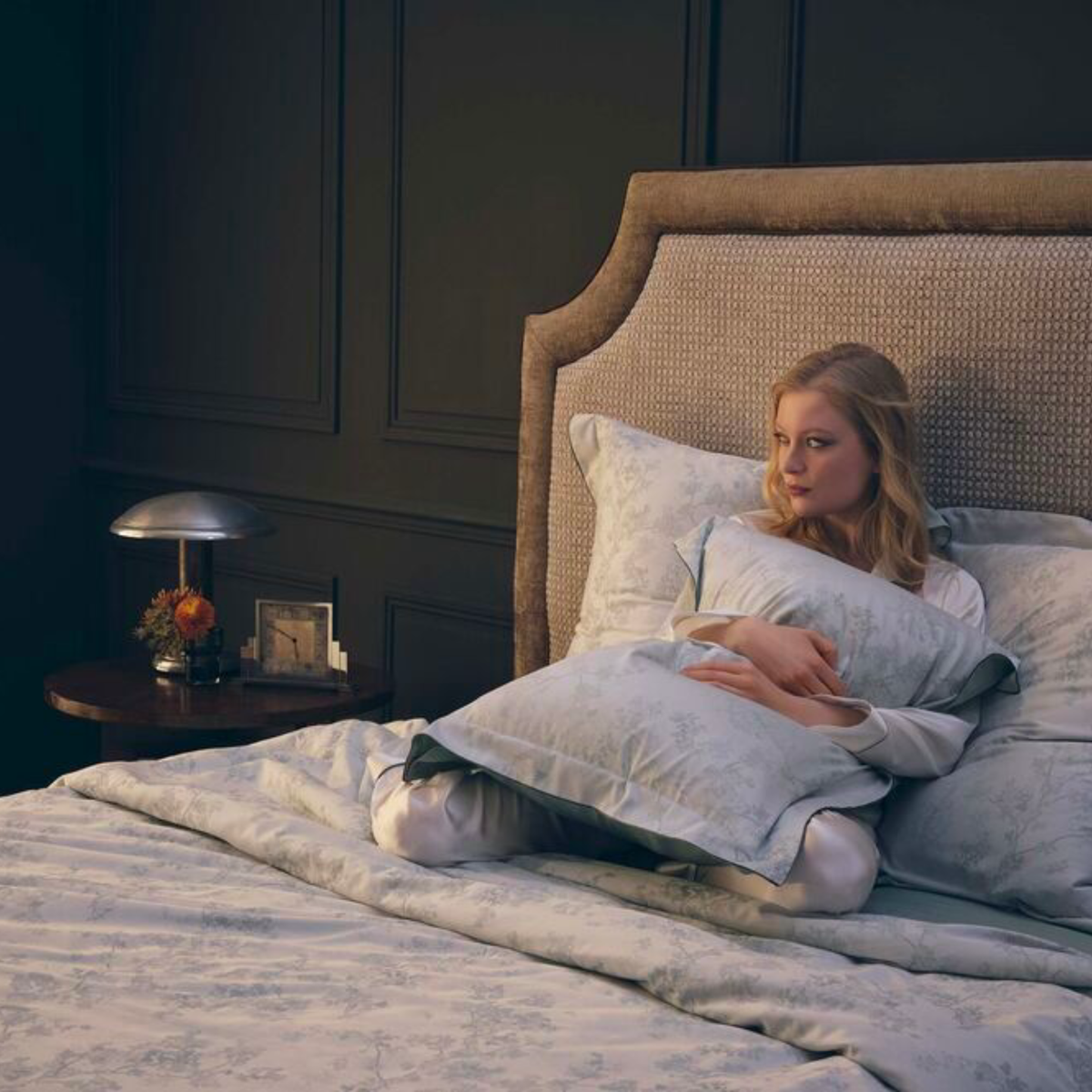 A woman sits on a bed with Yves Delorme Muses Bedding, holding a pillow beside a nightstand and lamp in a dimly lit bedroom.