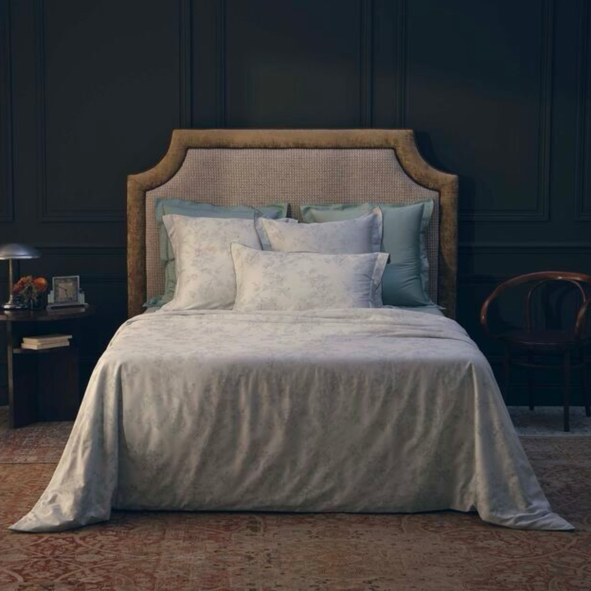 A neatly made bed with Yves Delorme Muses Bedding Collection, a wicker headboard, and two nightstands in a dark-walled bedroom.