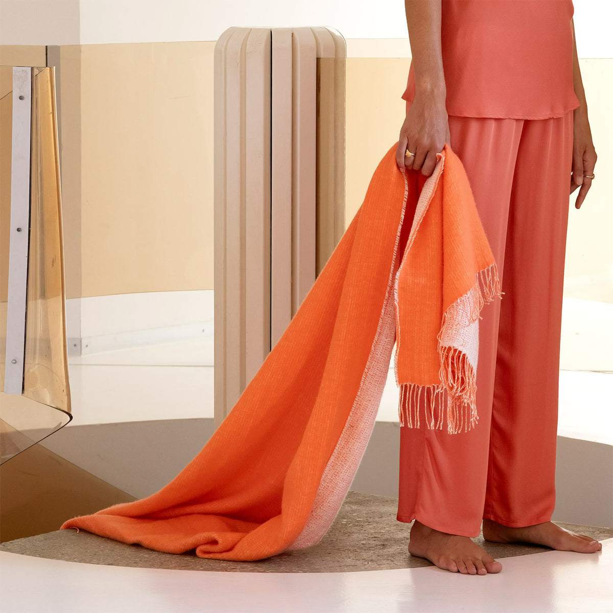 A person in coral pajamas stands barefoot, holding a Yves Delorme Mysore Throw in Corail color onto the floor