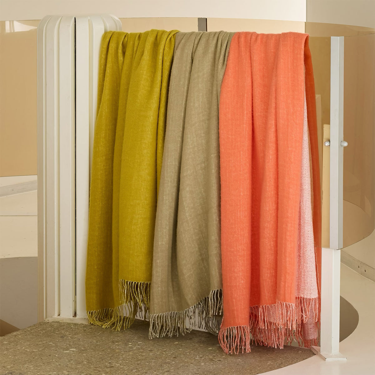 Three fringed Yves Delorme Mysore Throws hang over a white metal railing indoors