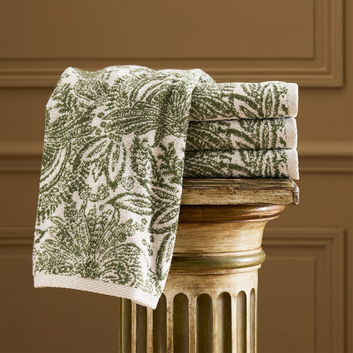 A stack of Yves Delorme Patio Bath Towels sits atop a gold fluted pedestal against a brown wall