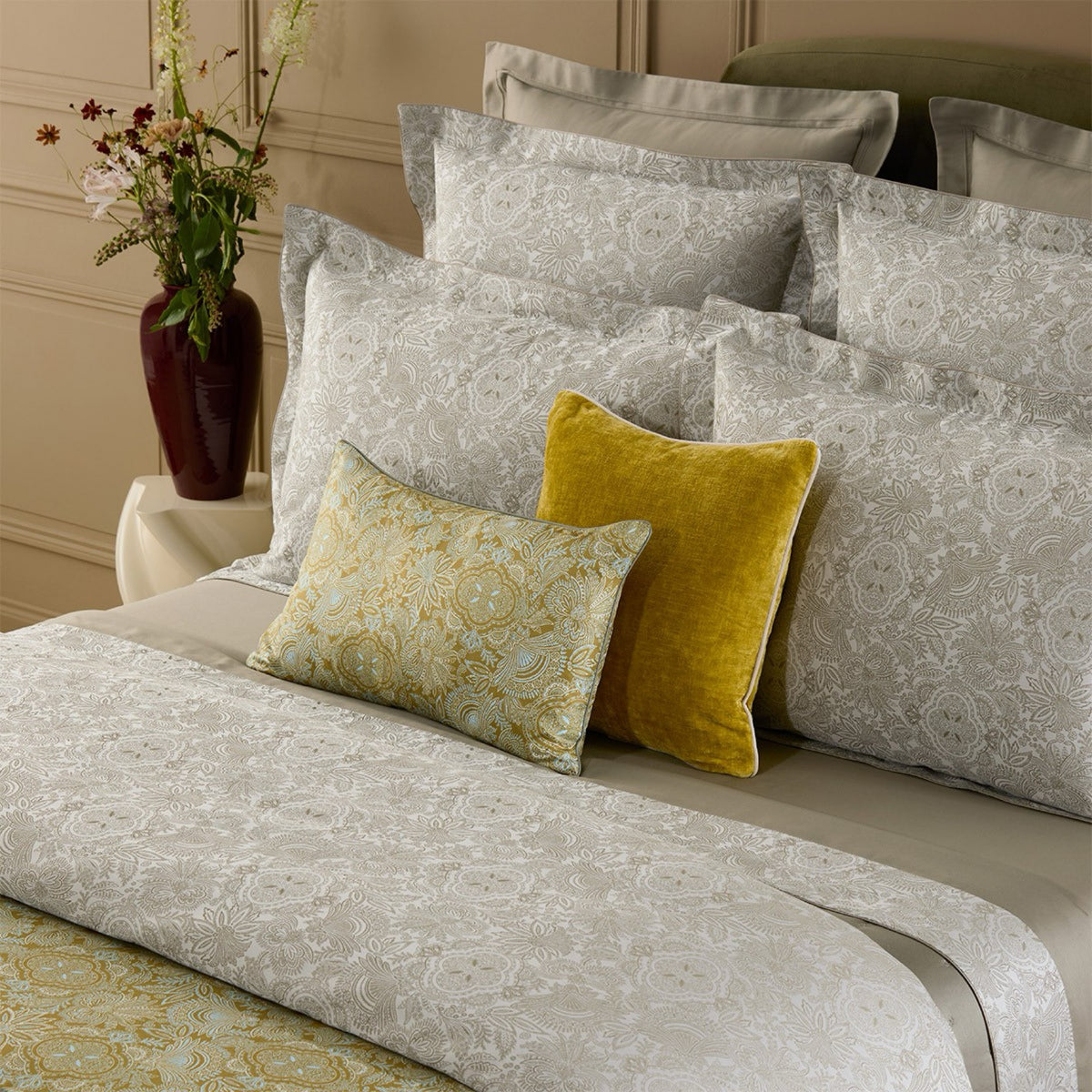 A neatly-made bed with Yves Delorme Patio Bedding, yellow accent pillows, and a vase of flowers on a side table.