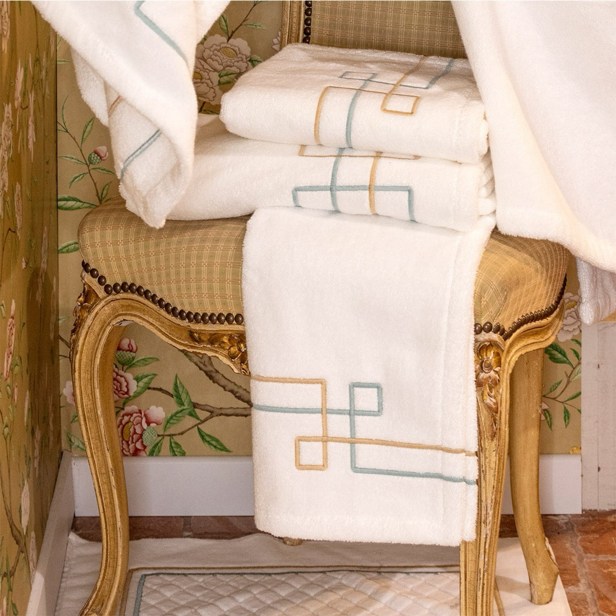 A stack of Yves Delorme Villa Bath Collection sitting on an ornate chair with a floral wallpaper background