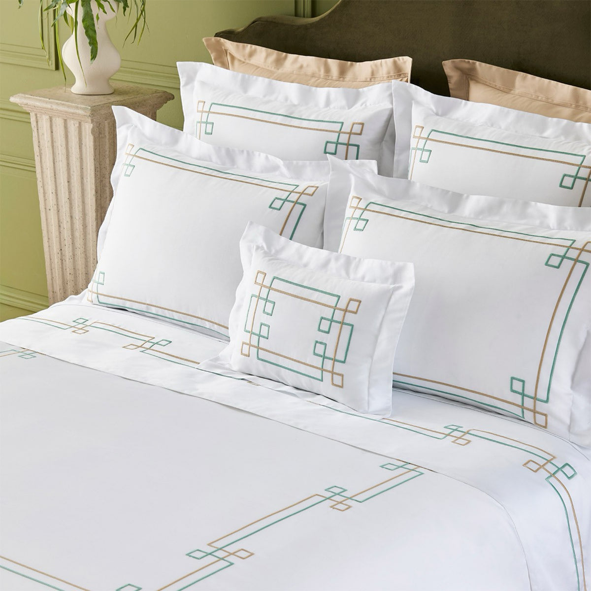 A neatly made bed with white linens from the Yves Delorme Villa Bedding, featuring a geometric green and gold pattern