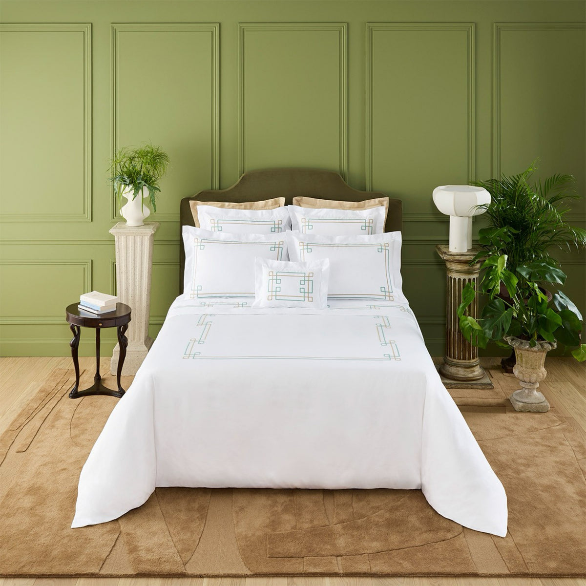 A neatly made bed with Yves Delorme Villa Bedding in white organic cotton satin, set in a green-walled room with plants