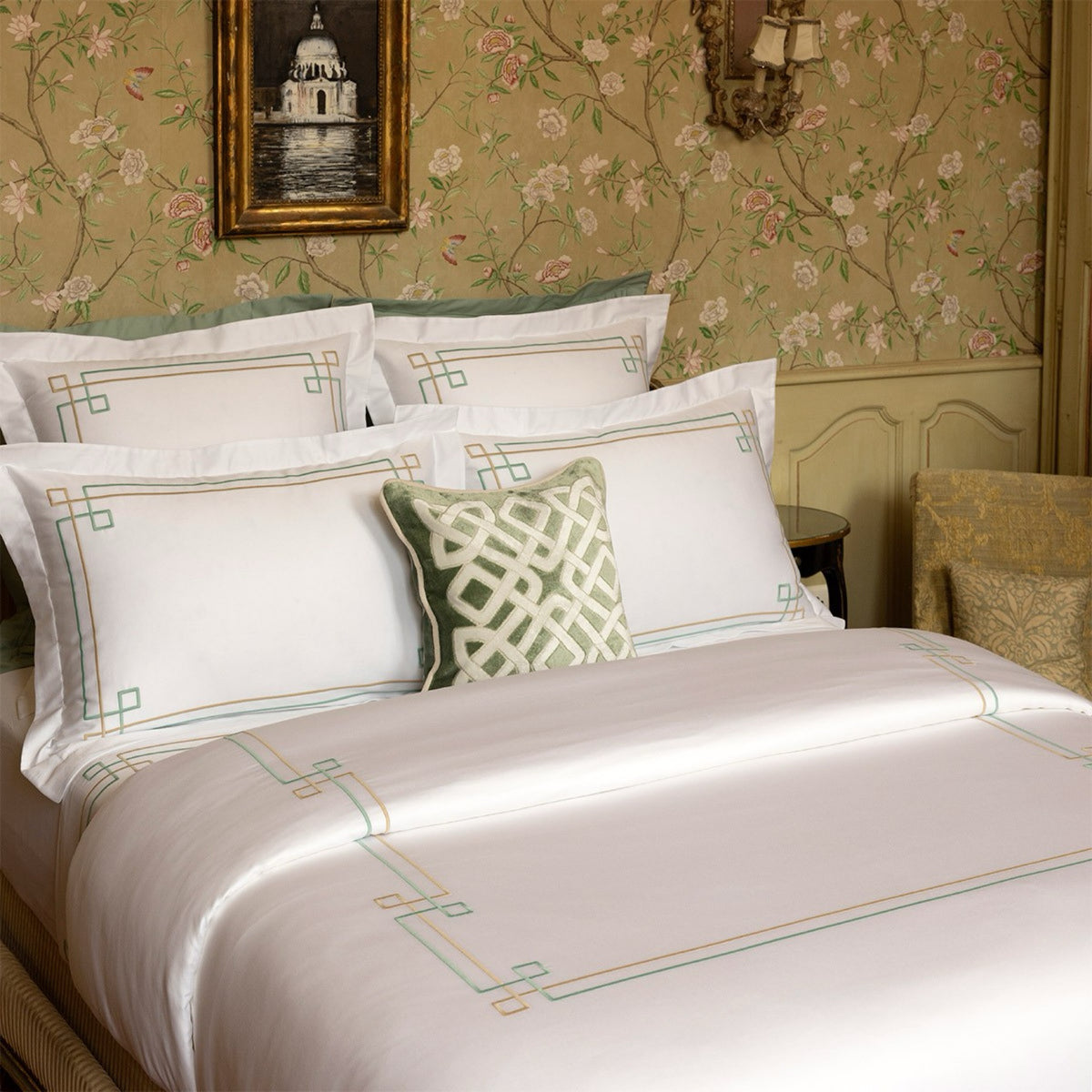 A neatly made bed with Yves Delorme Villa Bedding in a floral wallpapered bedroom