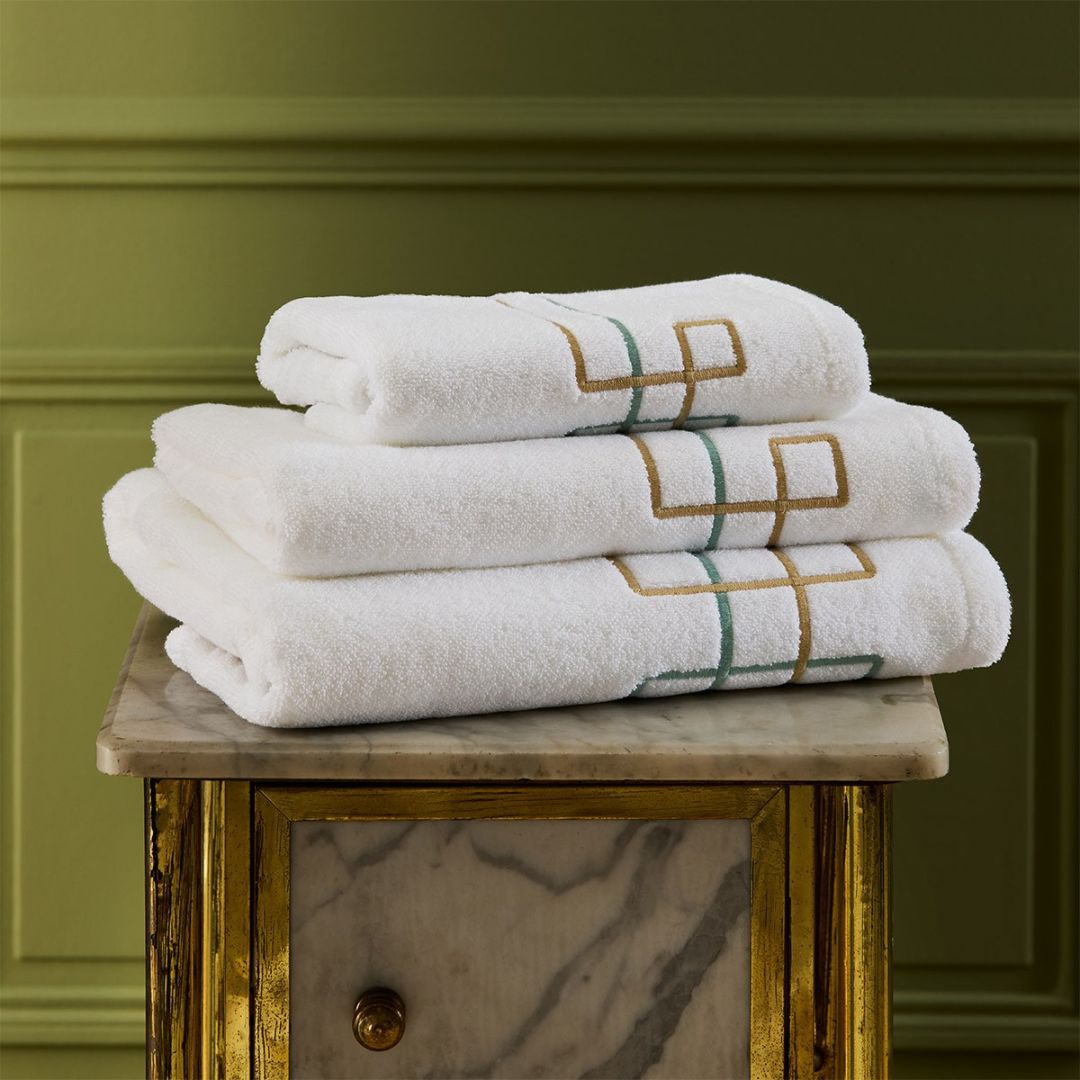 Three folded organic cotton bath towels with geometric embroidery are stacked on a marble-topped, gold-trimmed cabinet.