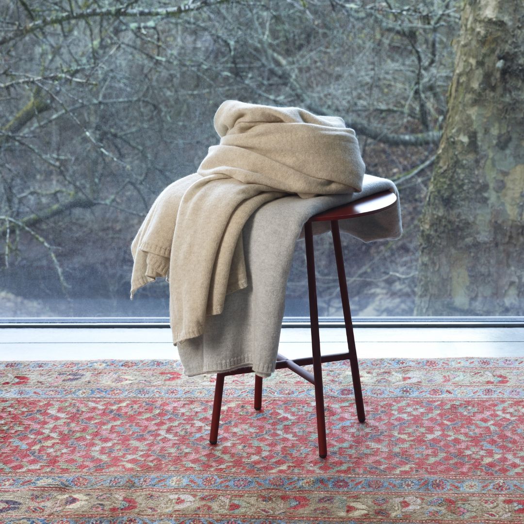 Two folded, luxury cashmere throws in neutral tones draped over a small, three-legged wooden stool.