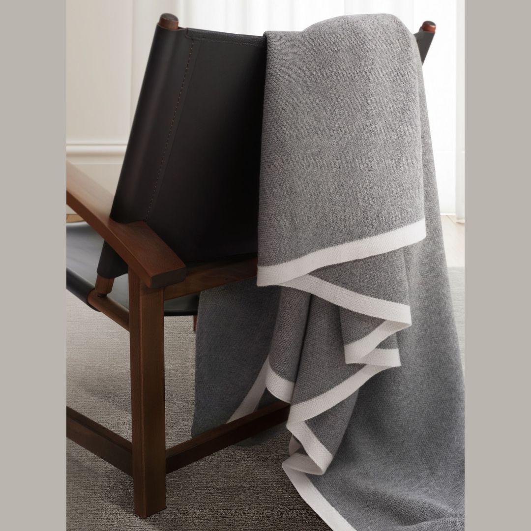 A gray cashmere silk throw with a white border draped over a dark wood and leather chair.
