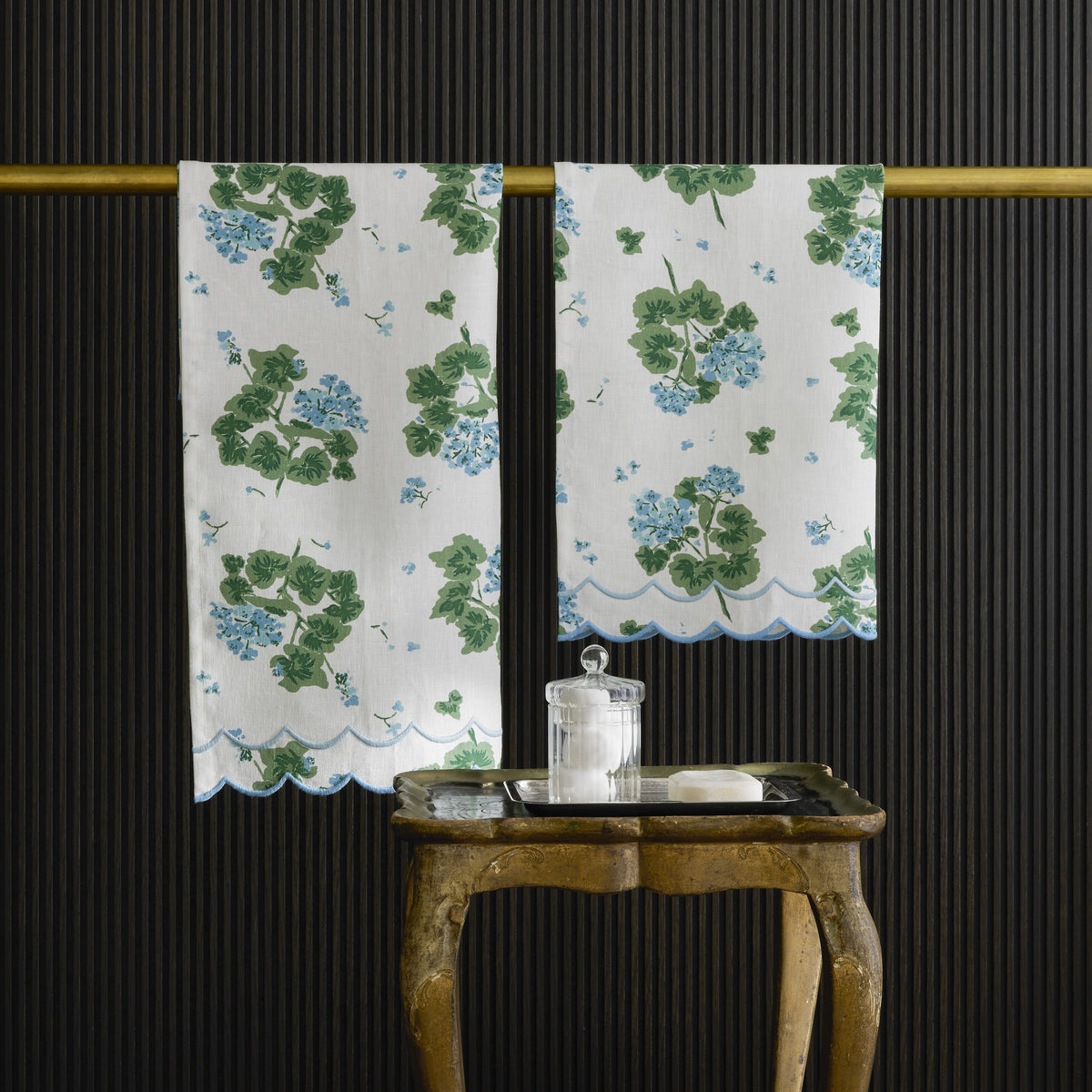 Floral-patterned guest towels hanging on a rod with a wooden table and decorative jar in the foreground against a black textured wall.