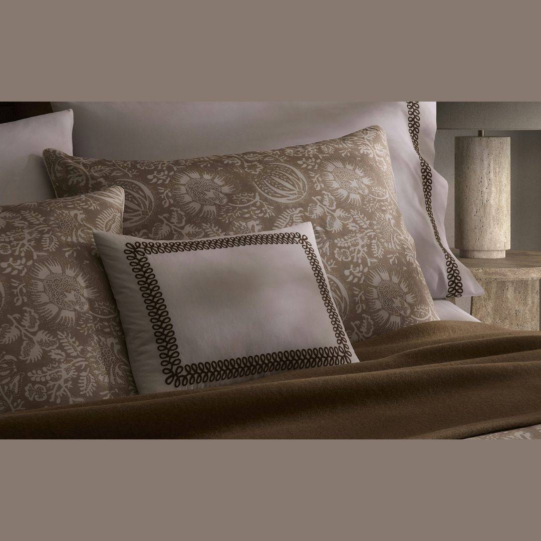 A bed with patterned pillows, a merino wool blanket, and a beige lamp on a nightstand in the background.