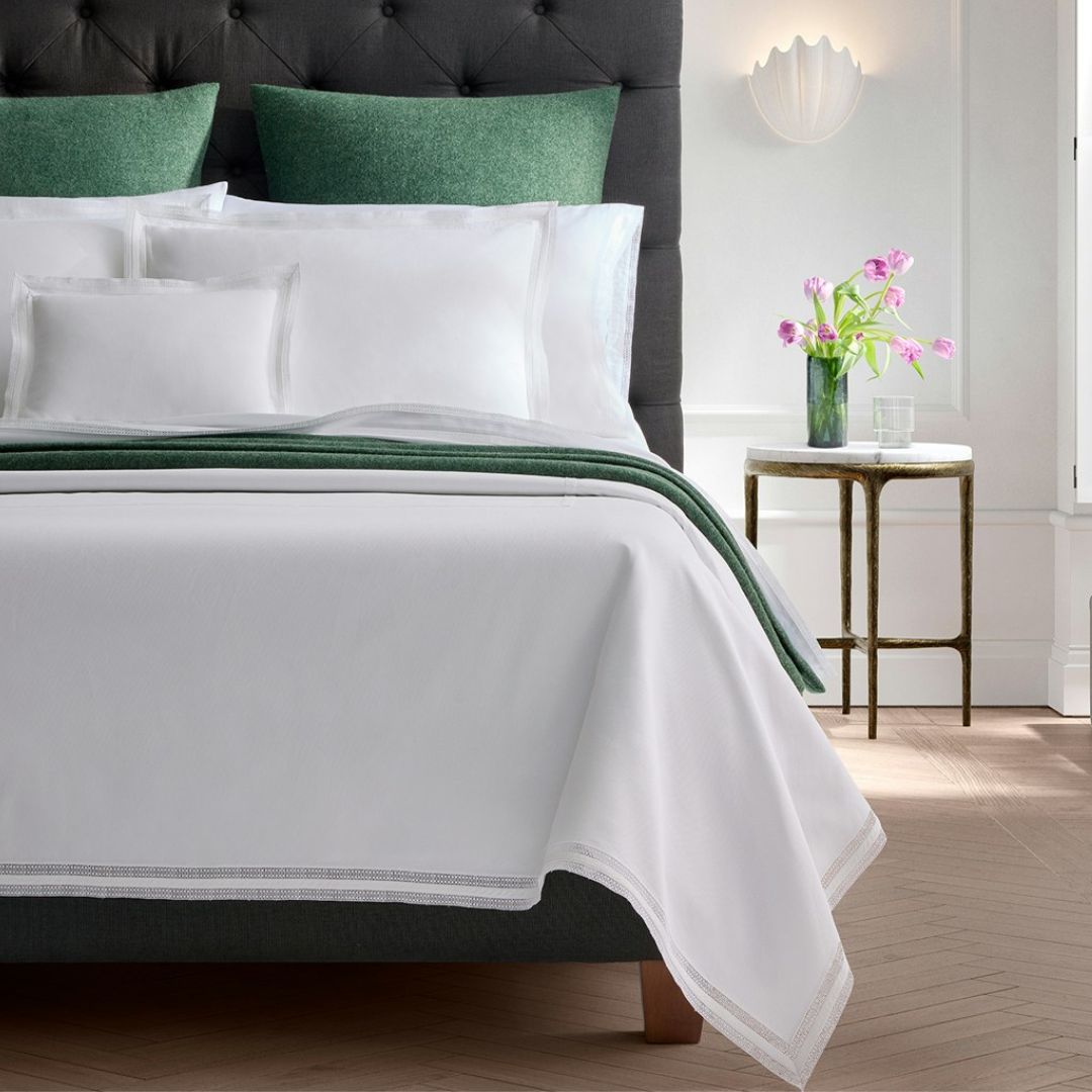 A neatly made bed with white linens, green pillows, and a Matouk Cosmo Blanket next to a round table with flowers.