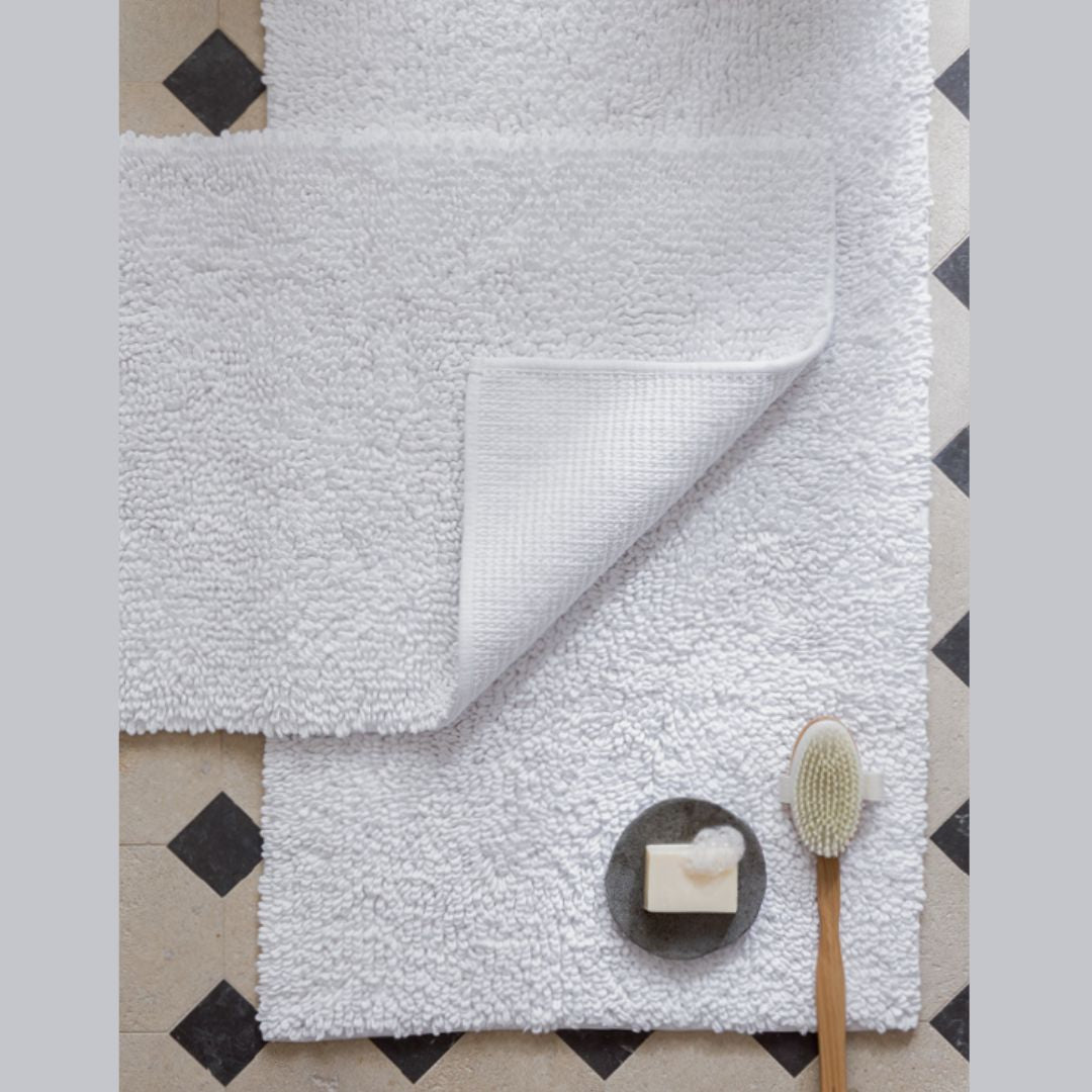 White Matouk Costas Bath Rug with one corner folded, beside a wooden bath brush and soap dish, on tile floor.