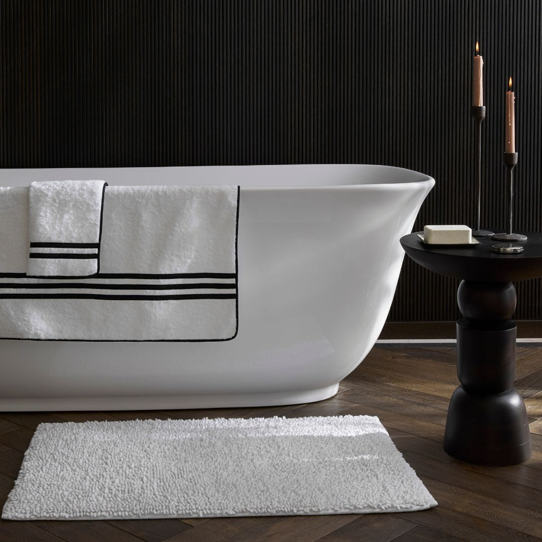 A white bathtub with striped towels, a Matouk Costas Bath Rug, and a small table with candles, soap, and a soap dish.