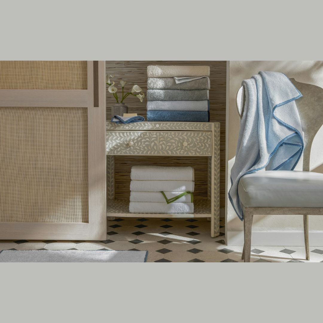 A stack of folded Enzo Bath Towels sits on a decorative table, with more below and one monogram draped over a white chair.