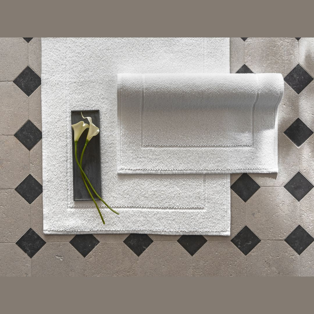 Two white Matouk Guesthouse textured bath rugs on tile, paired with a black tray holding two white calla lilies.