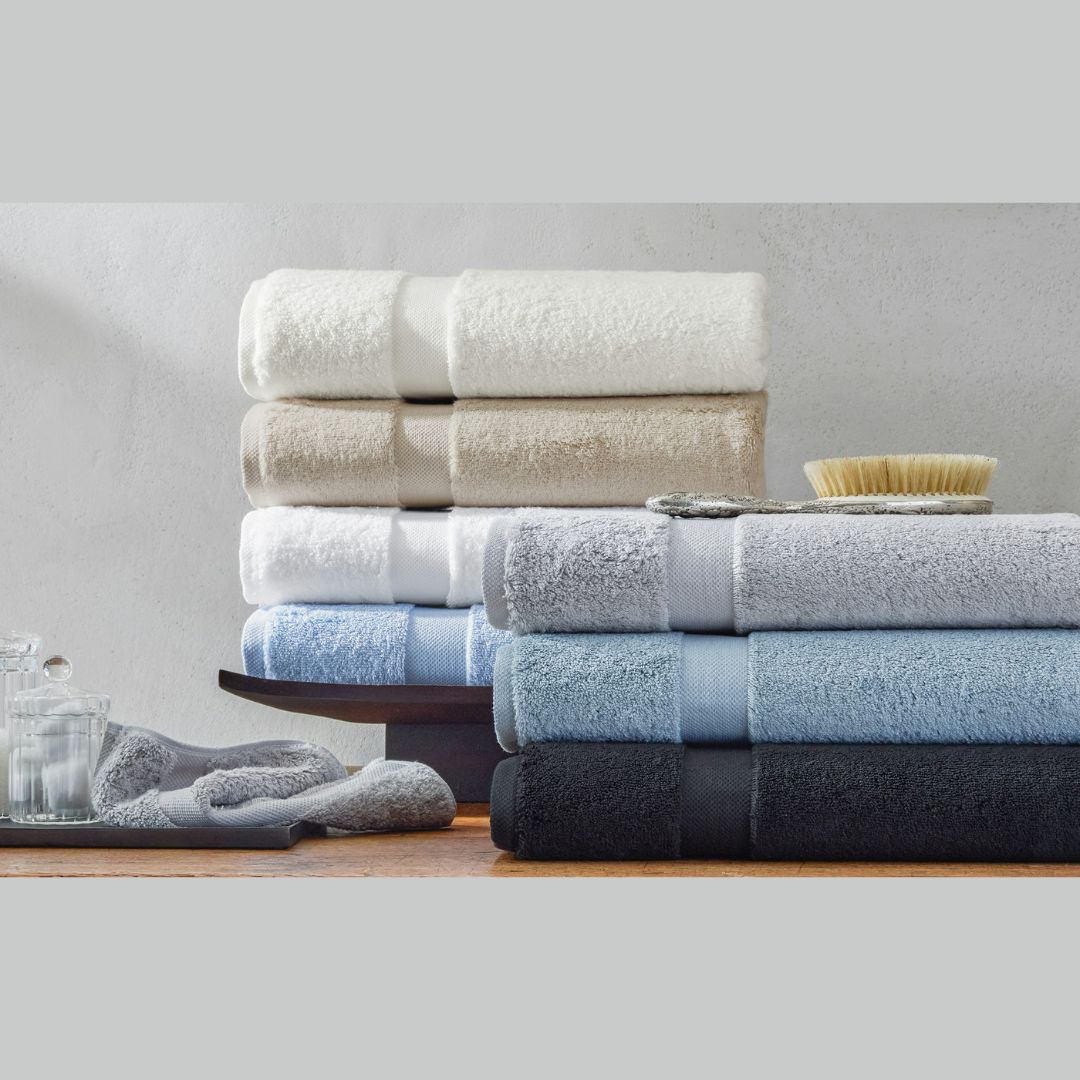 Plush cotton towels from the Matouk Lotus collection in soft hues are neatly stacked on a wooden surface.