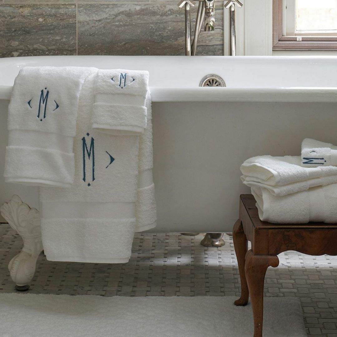 White Matouk Lotus collection towels hang on a clawfoot tub, with plush cotton towels stacked on a wooden stool nearby.