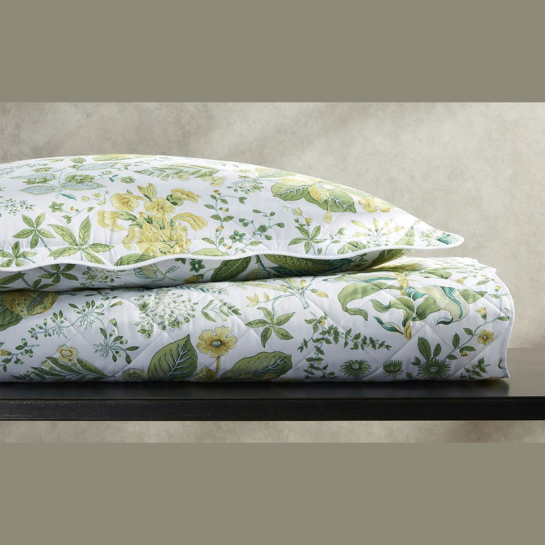 A folded comforter and pillow in Schumacher Matouk botanical print rest on a dark surface against a neutral wall.