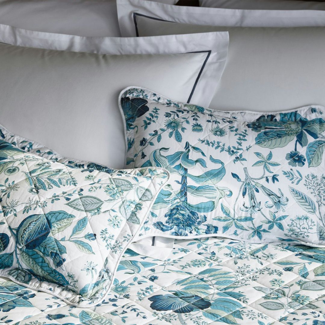 White bedding with blue piping is layered with Schumacher bedding and floral pillows in blue and green tones.