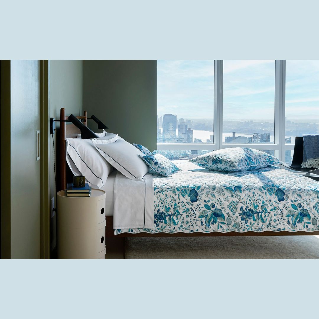 A neatly made bed with Schumacher bedding sits by a round nightstand; large windows reveal a city skyline.