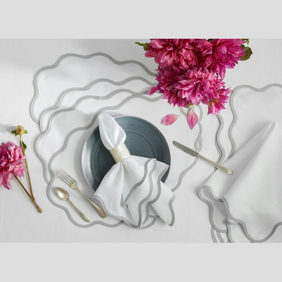 A table setting with linen placemats, cutlery, a plate, scallop edge napkins, and pink flowers arranged above.